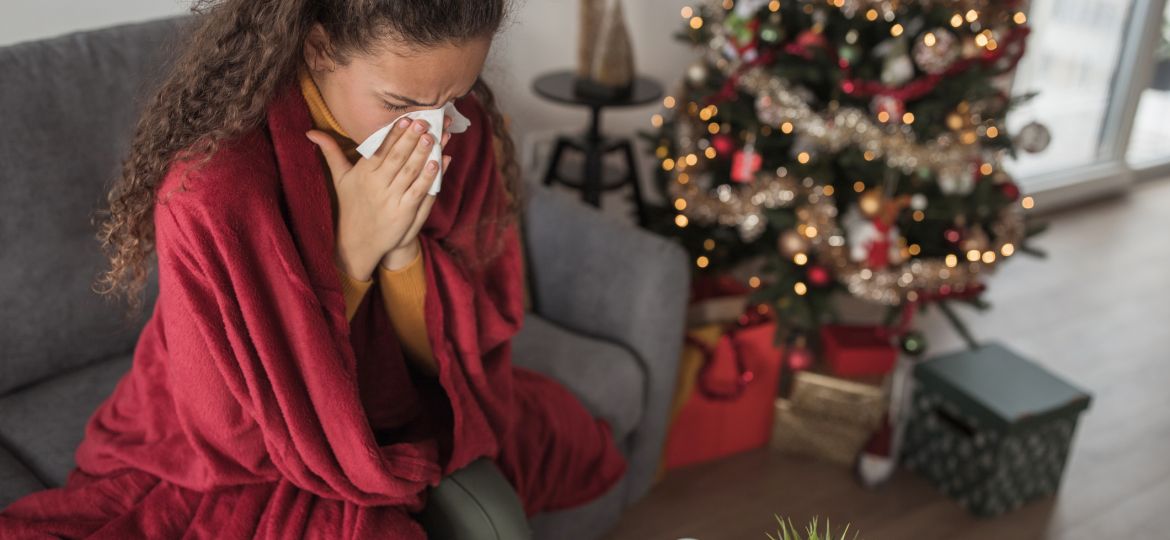 Young woman sitting by a Christmas tree covered with a blanket and blowing her nose