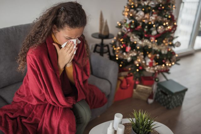 Young woman sitting by a Christmas tree covered with a blanket and blowing her nose